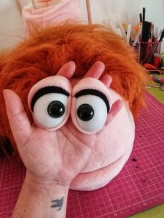 a hand is covering the face of a stuffed animal with large googly eyes and an orange wig