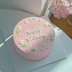 a pink birthday cake sitting on top of a white table next to a vase with flowers