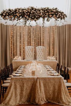 a table set up for a formal dinner with white chairs and flowers hanging from the ceiling