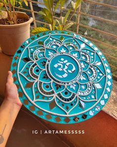 a person holding up a blue plate with the om shan symbol painted on it, in front of a potted plant