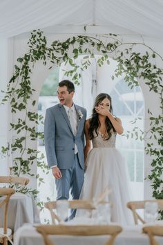 a bride and groom standing in front of a white tent with greenery on the walls