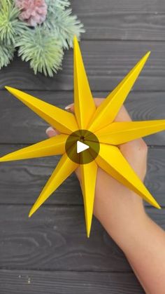 a person holding a yellow origami star on top of a wooden table next to a potted plant