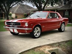 a red mustang sitting on top of a driveway next to a tree in front of a house