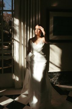 a woman in a white dress standing by a window with her hands on her head