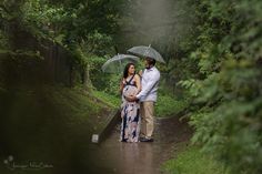 a pregnant couple standing under an umbrella on a path in the rain while holding hands and looking at each other