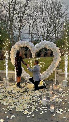 two people kneeling down in front of a heart shaped wreath with candles and flowers on the ground