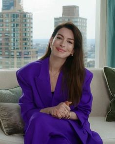 a woman sitting on top of a couch in front of a window with tall buildings behind her