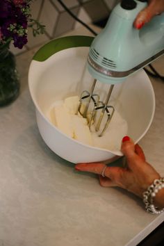 a woman mixing batter in a bowl with an electric mixer on the counter next to flowers