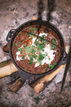 a pot filled with chili and sour cream on top of an open fire pit next to a pair of tongs