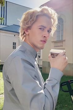 a young man holding a cup of coffee in front of a building with grass and trees