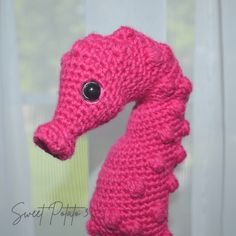 a pink knitted sea horse with black eyes and tail is shown in front of a window