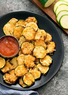 zucchini fritters with ketchup on a black plate next to sliced cucumbers