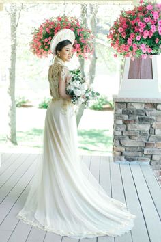 a woman in a white dress and hat standing on a porch with flowers behind her