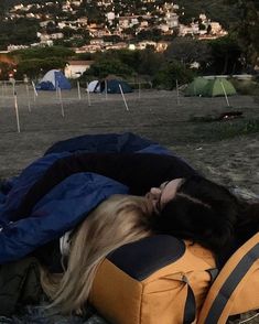 a woman laying on top of a yellow backpack next to a field with tents in the background