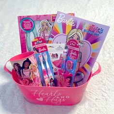 a pink bucket filled with lots of barbie dolls and books on top of a white blanket