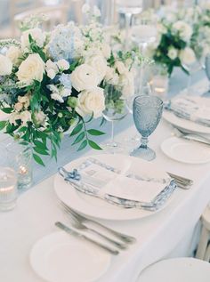 the table is set with white and blue flowers in vases, silverware, and napkins