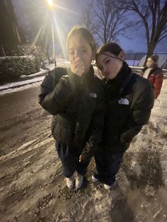 two people standing in the snow with their arms around each other and looking at the camera