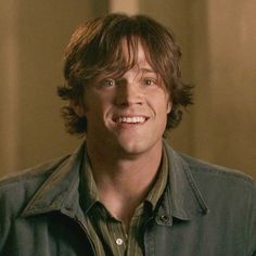 a young man with long hair smiling at the camera, wearing a green shirt and jacket