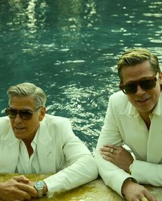 two men in white suits sitting next to each other on a ledge near the water