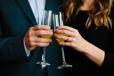 two people are holding wine glasses with yellow liquid in each hand and one person is wearing a black suit