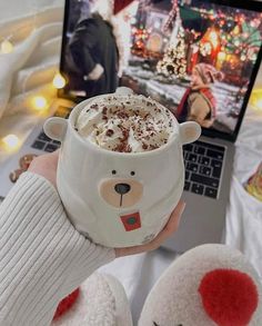 someone is holding a cup of hot chocolate in front of a laptop with a christmas scene on the screen