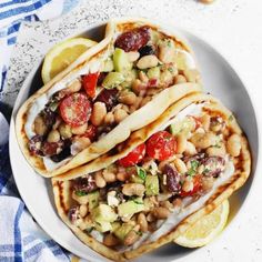 three tacos with beans, tomatoes, cucumbers and lemon wedges on a white plate