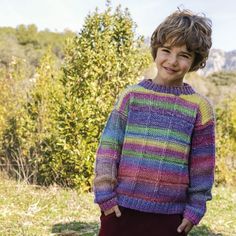 a young boy wearing a multicolored knitted sweater standing in front of some trees