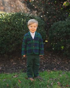 a young boy standing in the grass wearing a green and blue plaid jacket with white shirt