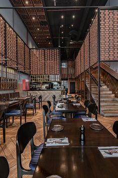 an empty restaurant with wooden tables and blue chairs in front of a brick wall that has stairs leading up to the second floor