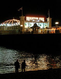 two people are standing on the beach at night by the water's edge, with lights in the background