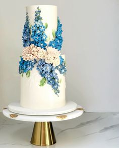 a white and blue wedding cake with flowers on the top is sitting on a gold stand