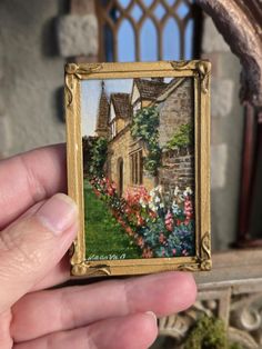 a hand holding up a miniature painting in front of a building with flowers growing on it