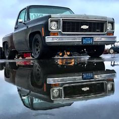 an old car is parked on the water with its reflection in the wet pavement and it's hood up