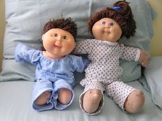 two dolls sitting on top of a bed next to each other, one is brown and the other is blue