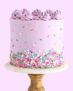 a pink cake with sprinkles and frosting on a white pedestal against a pink background