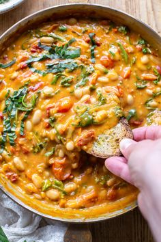 a hand dipping a piece of bread into a bowl of beans and spinach soup