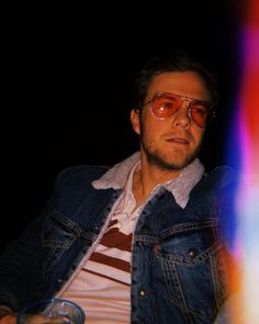 a man wearing glasses and a denim jacket sitting in front of a colorful light with his hand on his hip