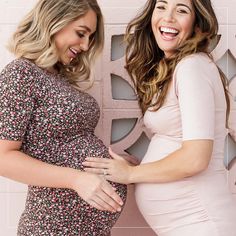 two pregnant women standing next to each other in front of a pink wall and smiling