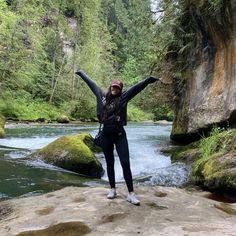 a woman standing on top of a rock in front of a river with her arms outstretched