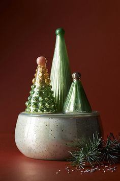 two green christmas trees sitting in a bowl on top of a red surface next to pine cones