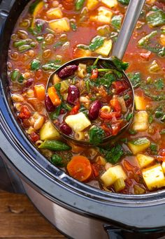 a ladle full of vegetable soup in a crock pot on top of a wooden table