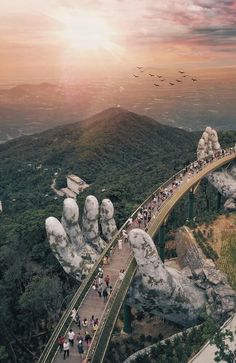 many people are walking on a bridge over some rocks and mountains with birds flying in the sky