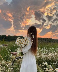 a woman in a white dress holding daisies standing next to a bike on a field