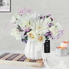 a white vase filled with lots of flowers on top of a wooden table next to a candle