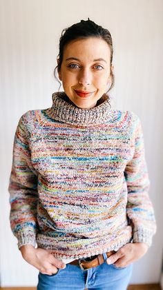 a woman standing in front of a white wall wearing a multicolored sweater and jeans