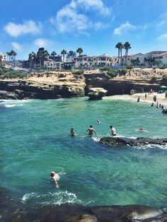 people are swimming in the ocean near some rocks and buildings with palm trees behind them