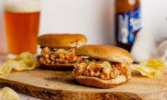 two pulled pork sandwiches on a cutting board with chips and beer in the back ground