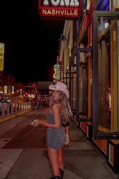 a woman standing on the side walk in front of a store at night with neon signs above her