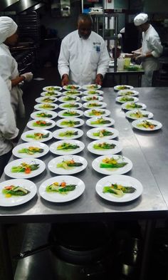 chefs preparing food in a commercial kitchen with many plates on the table and two men standing behind them