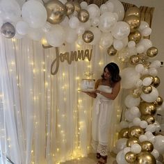 a woman holding a cake in front of a white backdrop with gold and silver balloons